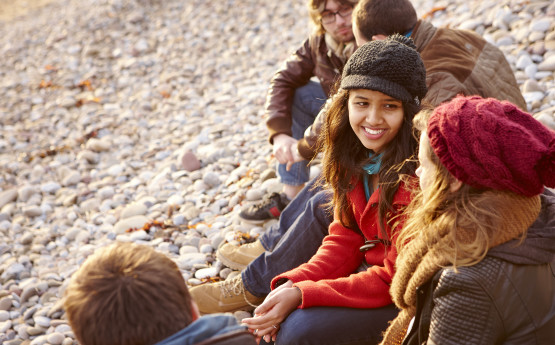 Four students sitting on a pebble beach, talking to each other. 