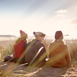 Three students sitting on the sand dunes of a beach talking and looking out to sea
